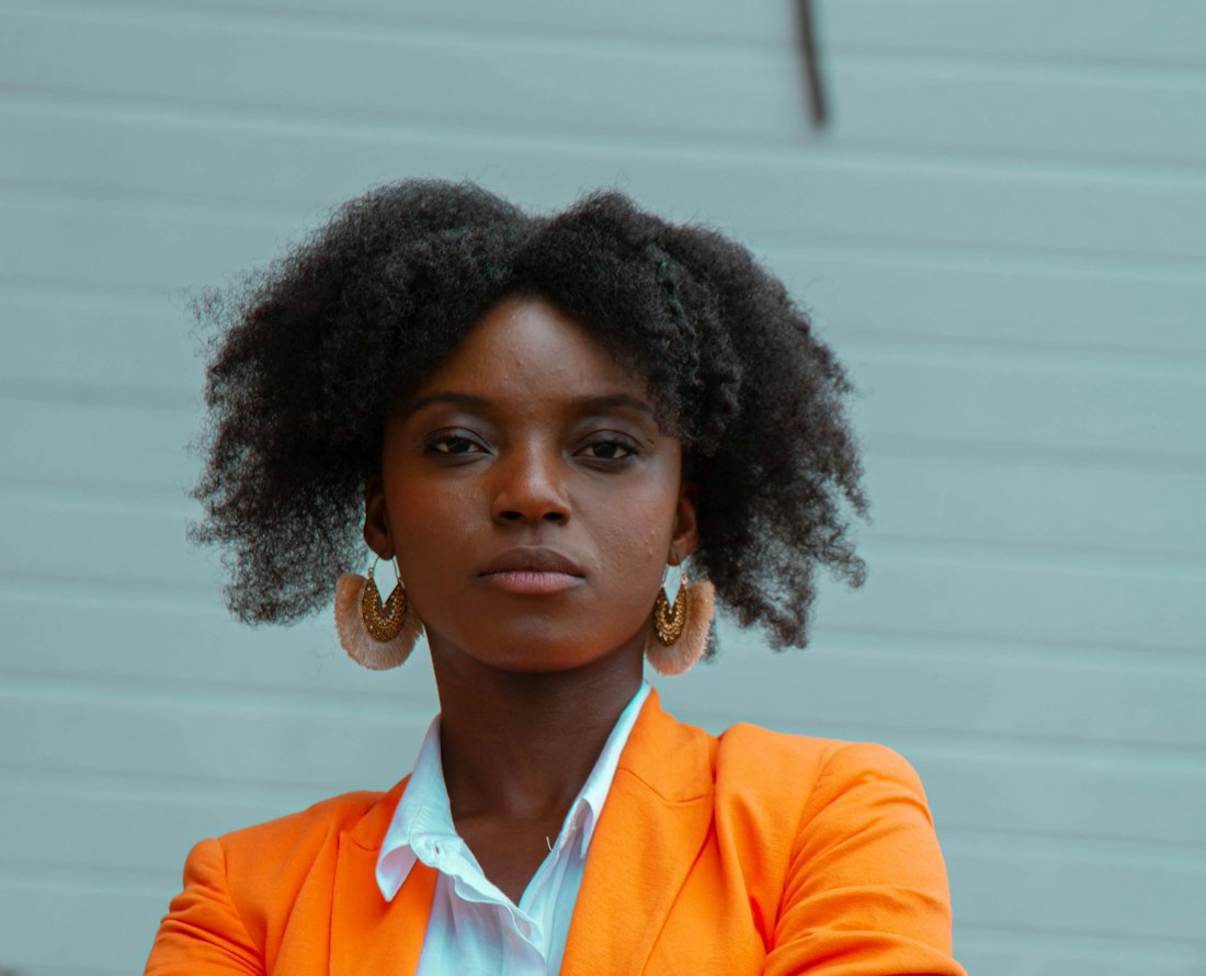 Conseillère en communication en blazer orange, bras croisés, devant un mur turquoise